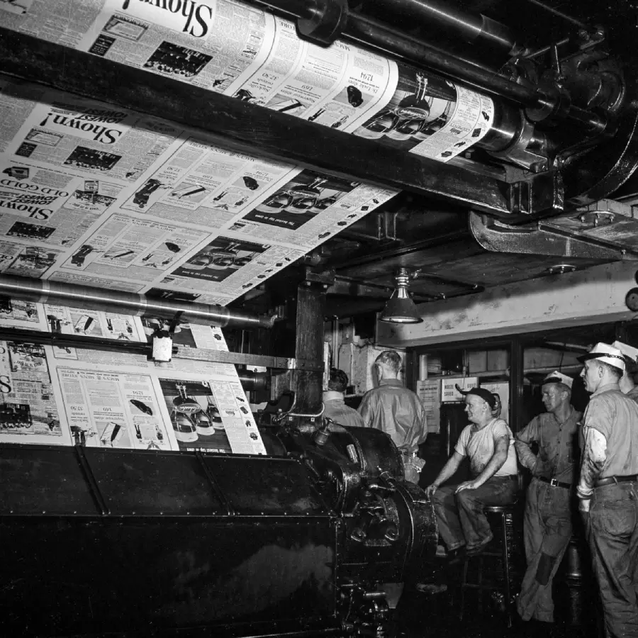 Men watch a large printing press in action as freshly printed newspapers move through the machine. The scene appears to be inside an industrial print shop.
