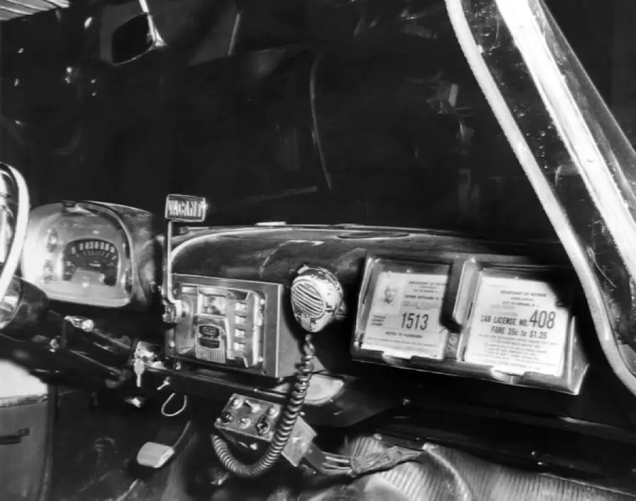 Black and white photo of a vintage car dashboard featuring a radio, microphone, speedometer, and two framed taxi license documents.