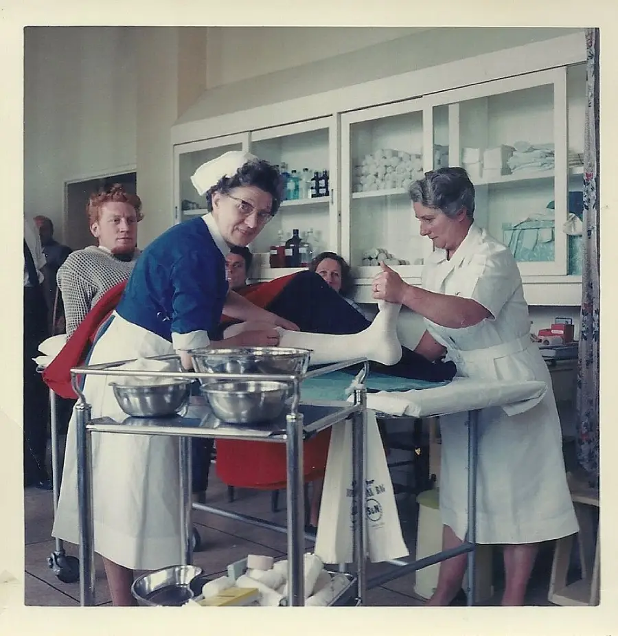 Two nurses in white uniforms apply a cast to a patient’s leg in a medical room, with shelves of supplies in the background. A man and another woman observe the procedure. Metal bowls and medical tools are on a cart nearby.