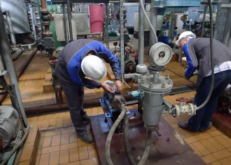 Two workers in safety helmets and uniforms repair machinery in an industrial facility. Pipes, gauges, and large equipment surround them on a tiled floor. They are focused on maintenance tasks in a mechanical setting.