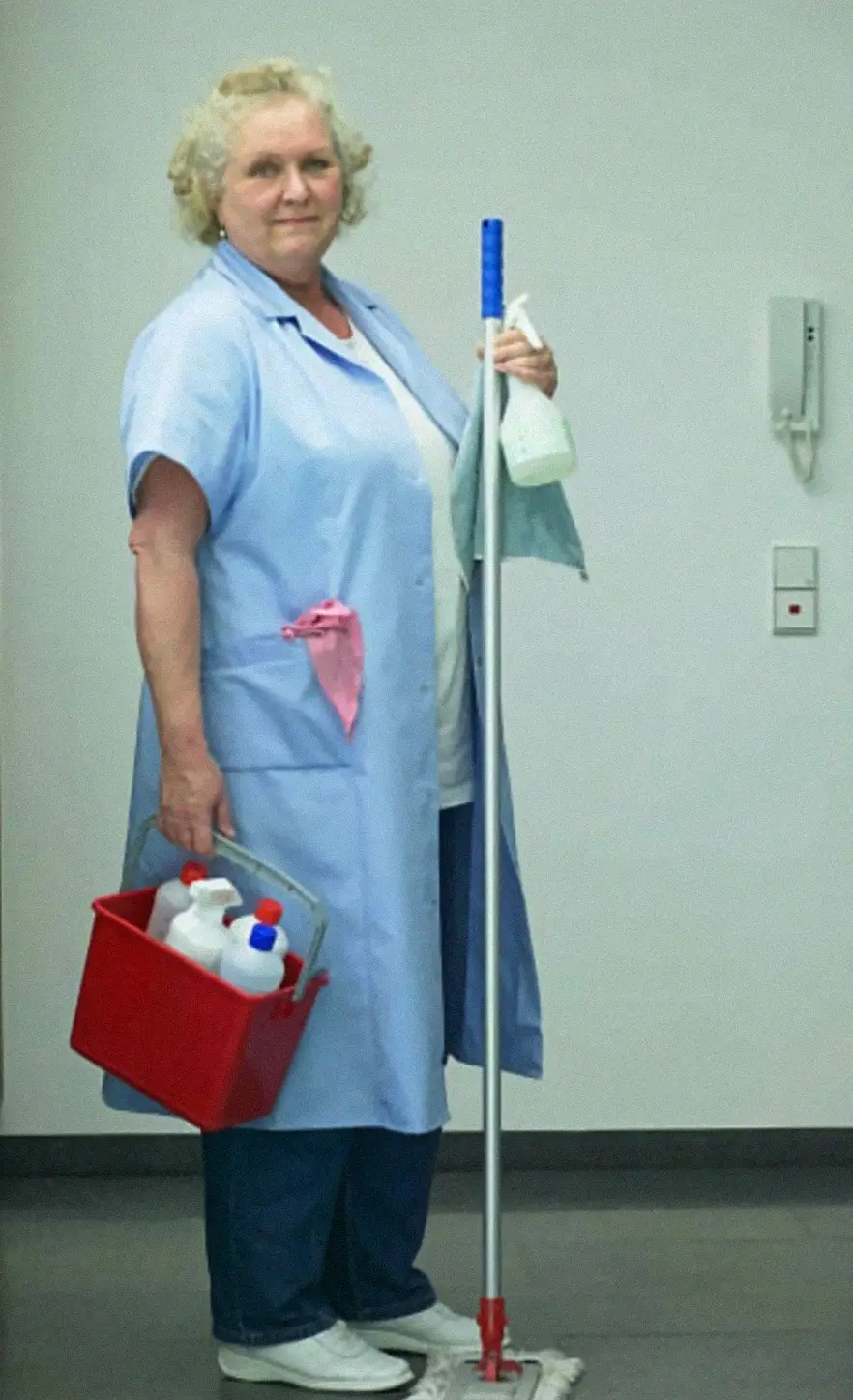 A woman in a blue uniform holds a mop, a red cleaning bucket with bottles, and a spray bottle, standing in a clean hallway with a white wall and a wall-mounted intercom.