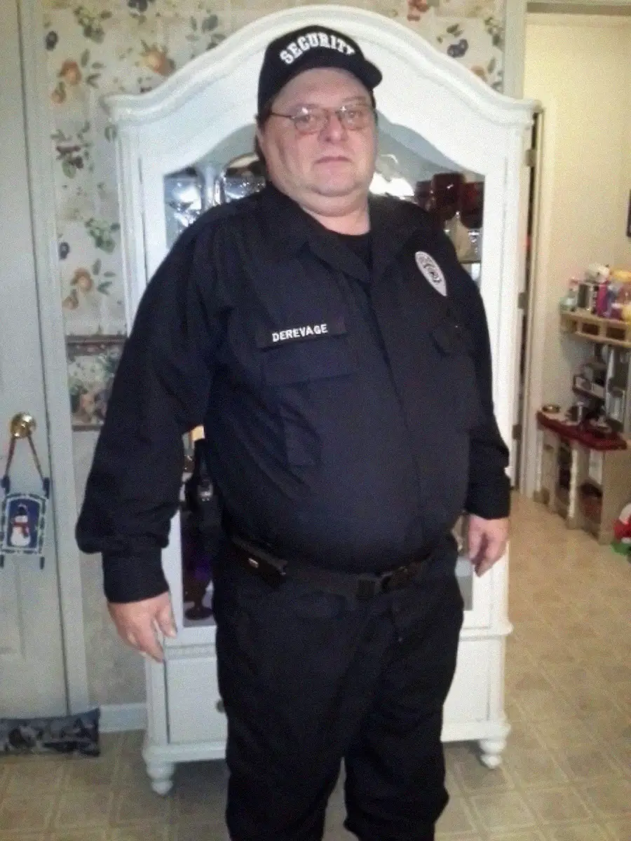 A man in a black security uniform and cap stands indoors in front of a white dresser. The uniform has a name tag that reads "Dreyage." The room has floral wallpaper and various items on shelves.