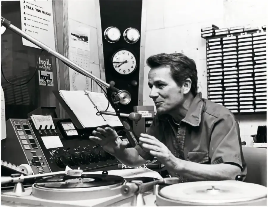 A man sits at a radio station desk, speaking into a microphone. There are turntables, control panels, and shelves with tapes behind him. A large clock and posted notes are visible on the wall.
