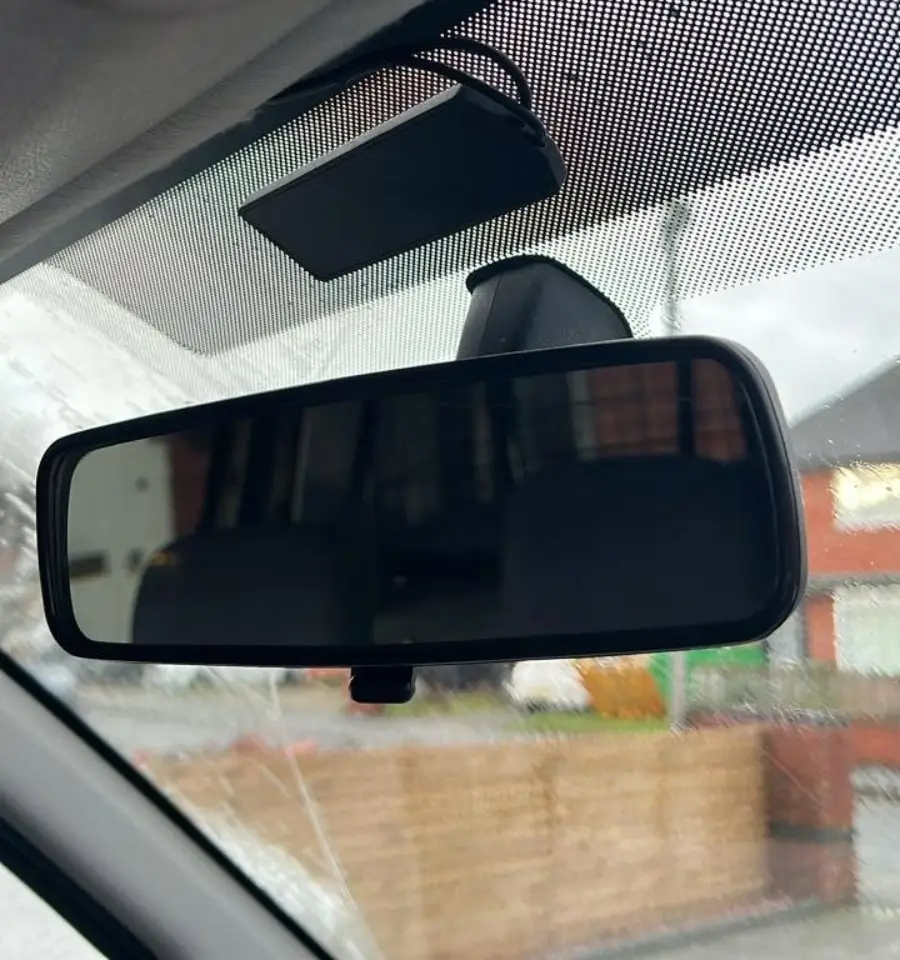 A car rearview mirror shows a clear reflection of the inside of the car and outside houses, with raindrops visible on the window, and a small device mounted above the mirror.