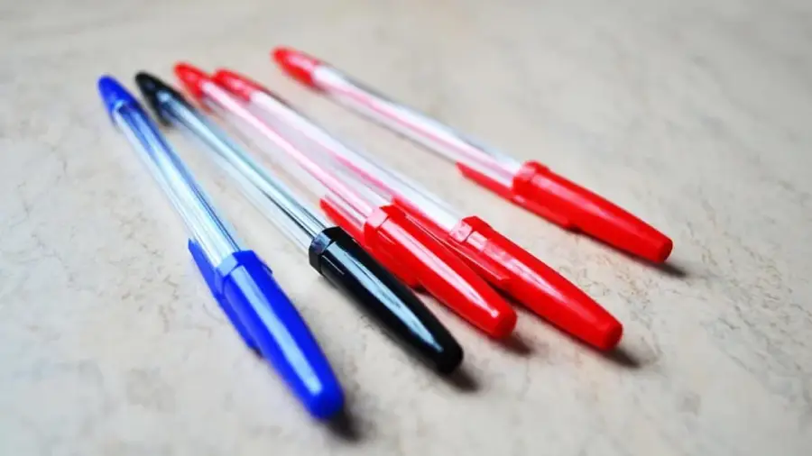 Five ballpoint pens with transparent barrels and colored caps—two blue, one black, and two red—are placed diagonally on a light, textured surface.