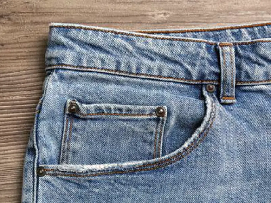 Close-up of the front pocket area of blue jeans, showing detailed stitching, a small coin pocket, and a brown wooden surface in the background.
