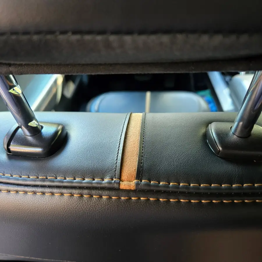 Close-up view of a car seat’s black leather headrest with tan stitching and a tan center stripe, showing the metal supports and stitching details.
