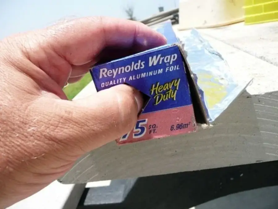 A hand presses a tab on the side of a Reynolds Wrap aluminum foil box, securing the roll in place while dispensing the foil.