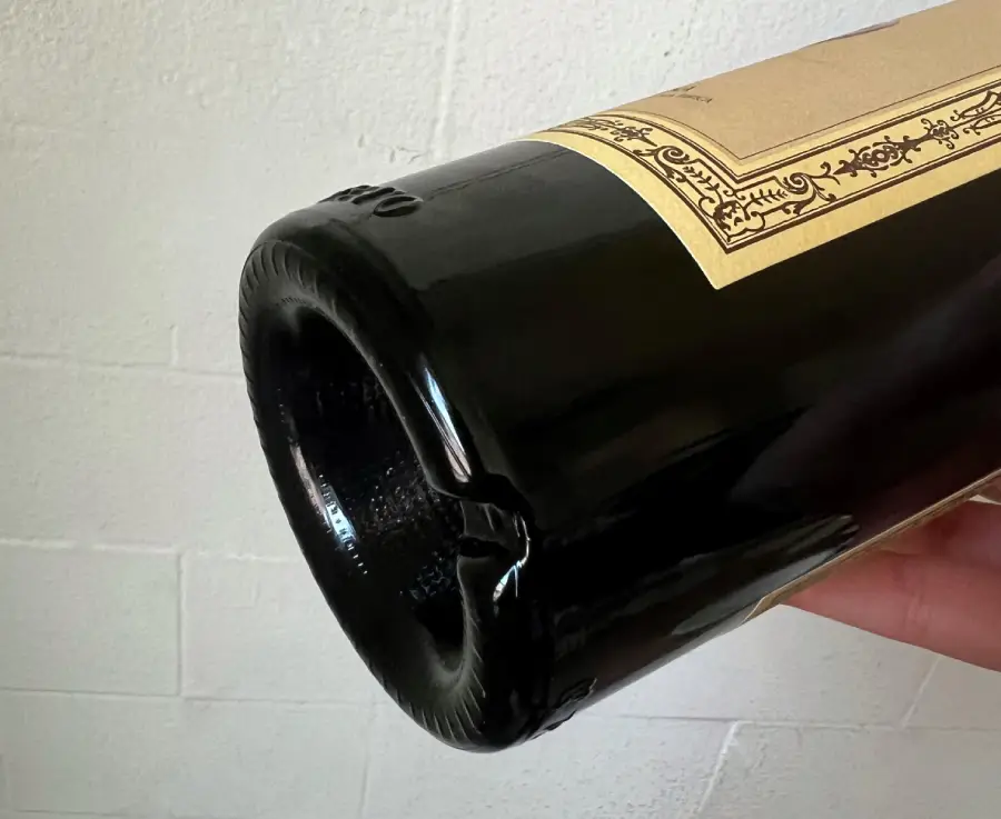 A close-up of the bottom of a dark glass wine bottle with a deep indentation, known as a punt, and a partial view of a beige label. The background is a white textured wall.