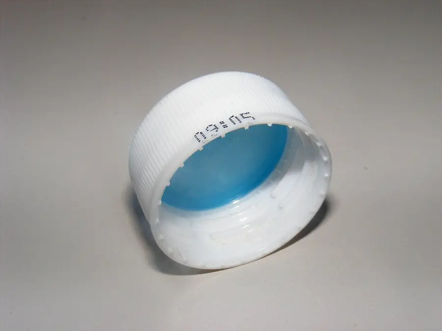 A close-up of a white plastic bottle cap with a blue inner seal, lying on its side on a plain, light-colored surface. Black printed numbers and letters are visible on the cap's outer edge.