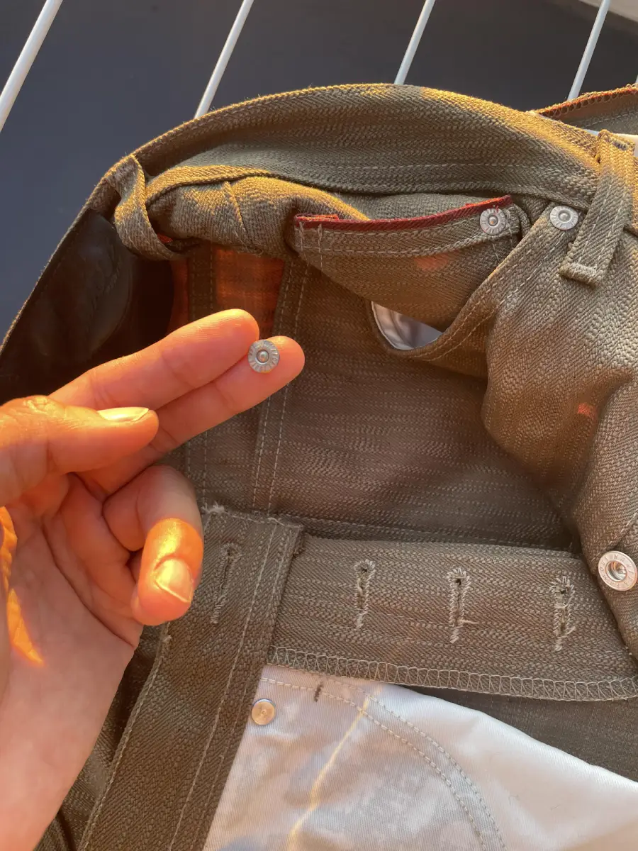 A hand holds a small button that has come off a pair of khaki pants, showing its detached position near the waistband and buttonhole area in warm sunlight.