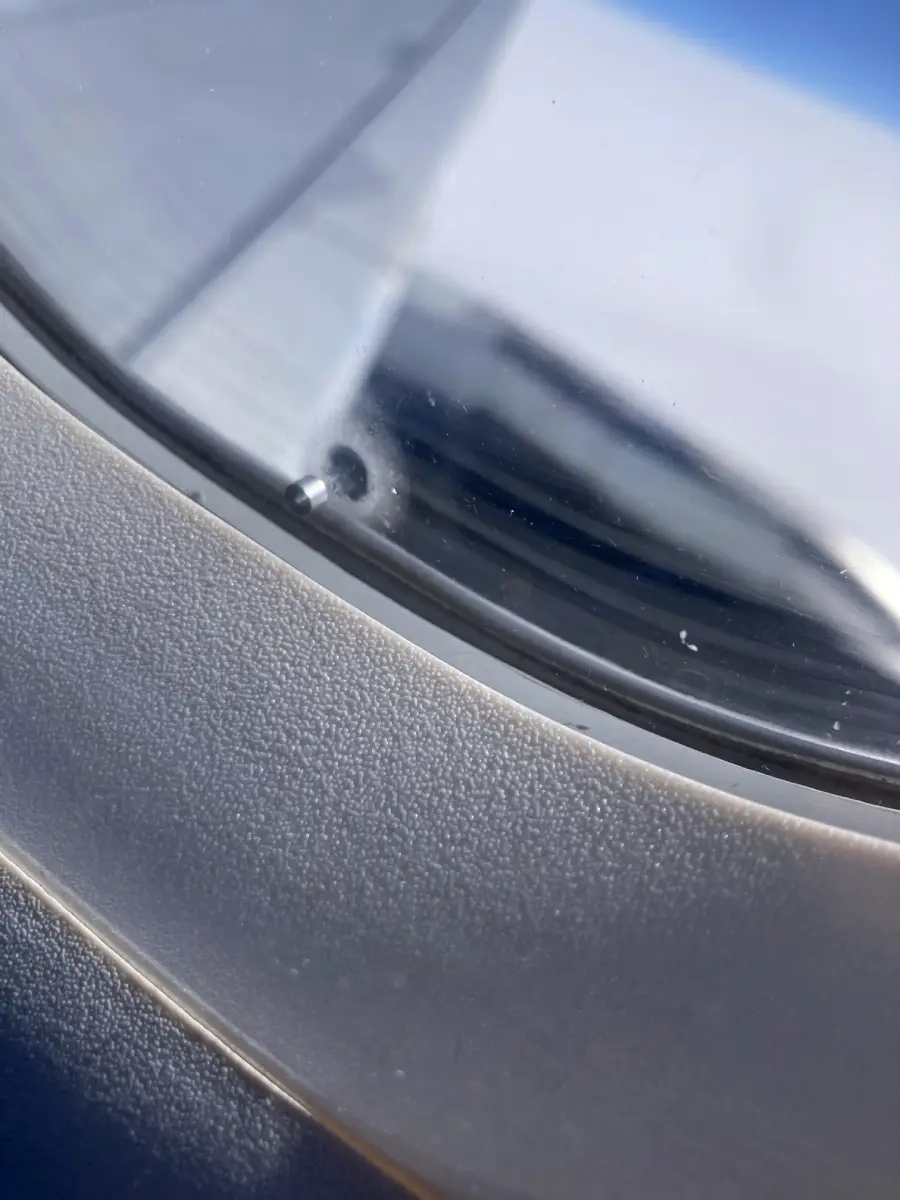 Close-up view of an airplane window showing the tiny breather hole in the middle, with the aircraft wing and bright sky visible outside.
