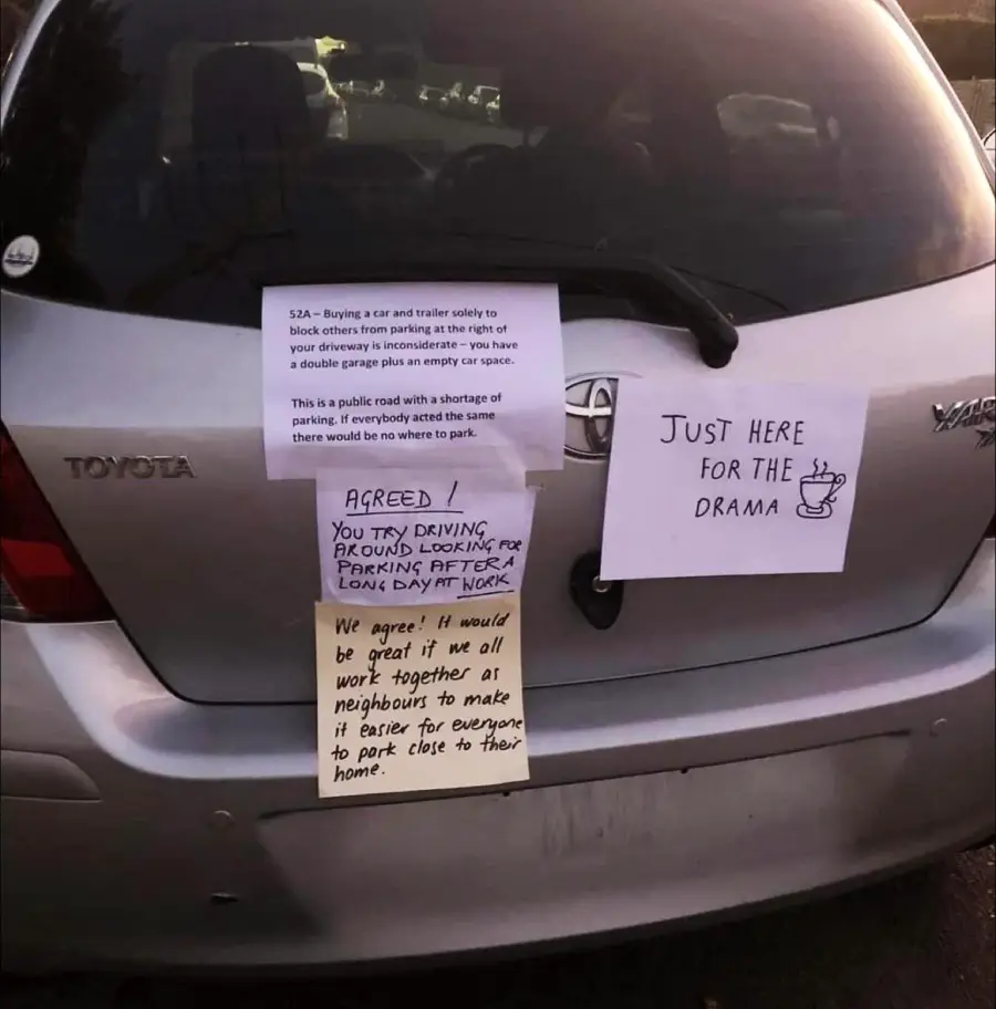 A silver Toyota car with several notes taped to its trunk, discussing parking issues. One says "Just here for the drama" with a coffee cup drawing; others debate double parking and cooperation among neighbors.
