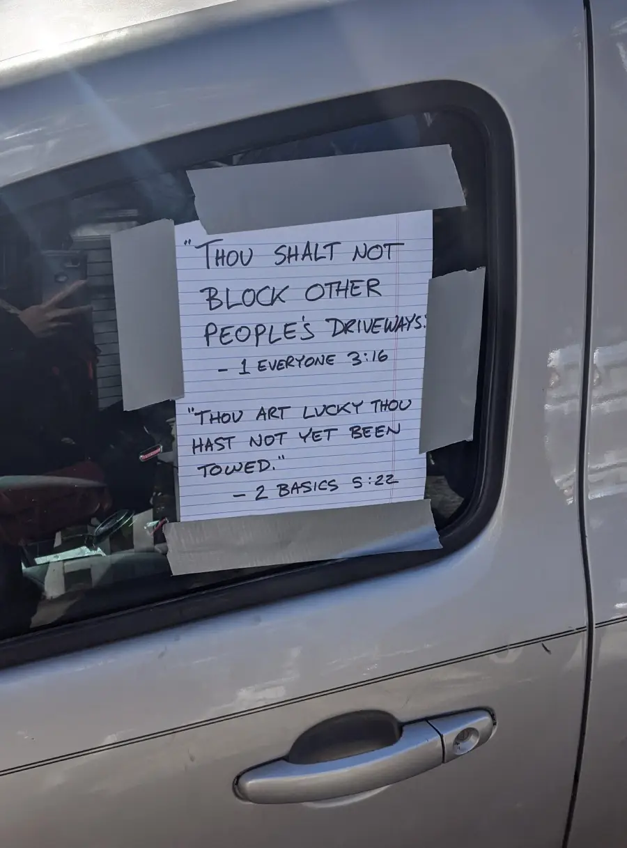 A handwritten sign taped to a car window reads: "'Thou shalt not block other people's driveways.' - Everyone 3:16. 'Thou art lucky thou hast not yet been towed.' - 2 Basics 5:22.