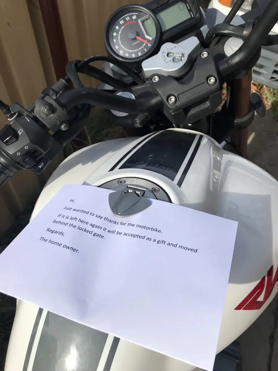 A close-up of a motorcycle's handlebars and fuel tank with a typed note placed on it, warning that if the bike is left again, it will be considered a gift and moved.