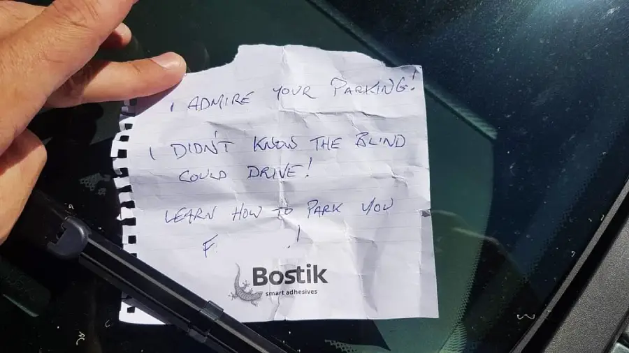 A handwritten note under a windshield wiper reads, "I admire your parking! Didn’t know the blind could drive! Learn how to park you f..." A hand is lifting the note.