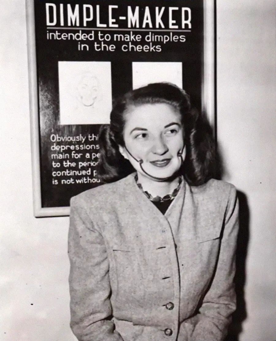 A woman wearing a device on her cheeks sits in front of a sign that reads “DIMPLE-MAKER intended to make dimples in the cheeks.” She is smiling and looking slightly to the side.