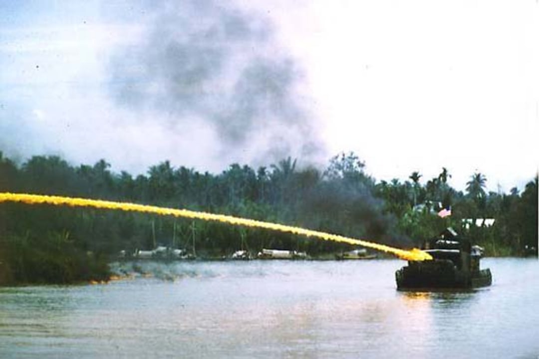 A military boat on a river shoots a stream of fire toward the riverbank, with smoke rising and palm trees in the background under a cloudy sky.