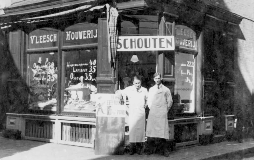 Two people in white aprons stand outside a corner butcher shop with large windows displaying signs and meats. The shop has the name “Schouten” above the door and Dutch words on the windows. The photo is black and white.