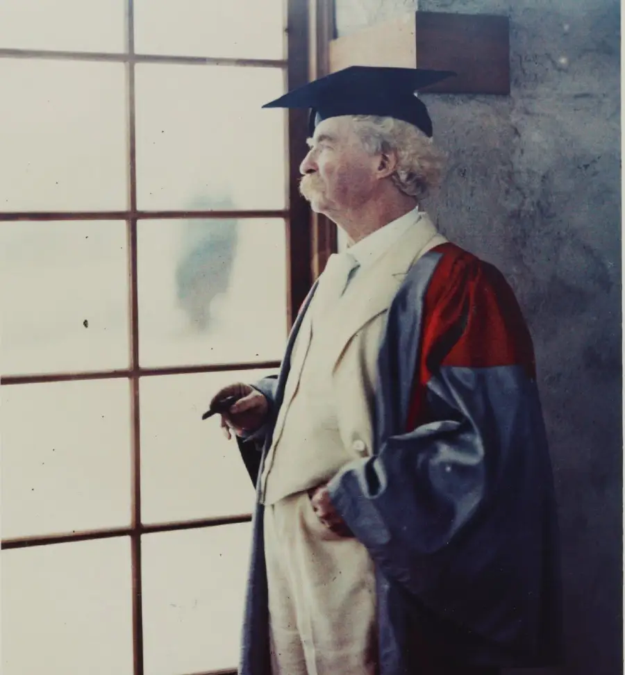 An older man with white hair and mustache, wearing a graduation cap and robe, stands indoors by a window, holding a cigar and looking outside.