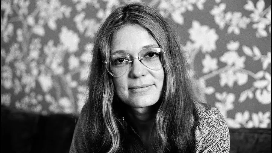 A woman with long hair and glasses looks at the camera, wearing a patterned top. The background features a floral wallpaper in black and white.
