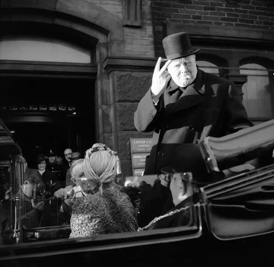 A man in a top hat and coat makes a gesture with his hand while standing in a car. Several people are seated with him, and onlookers watch from the background outside a brick building.