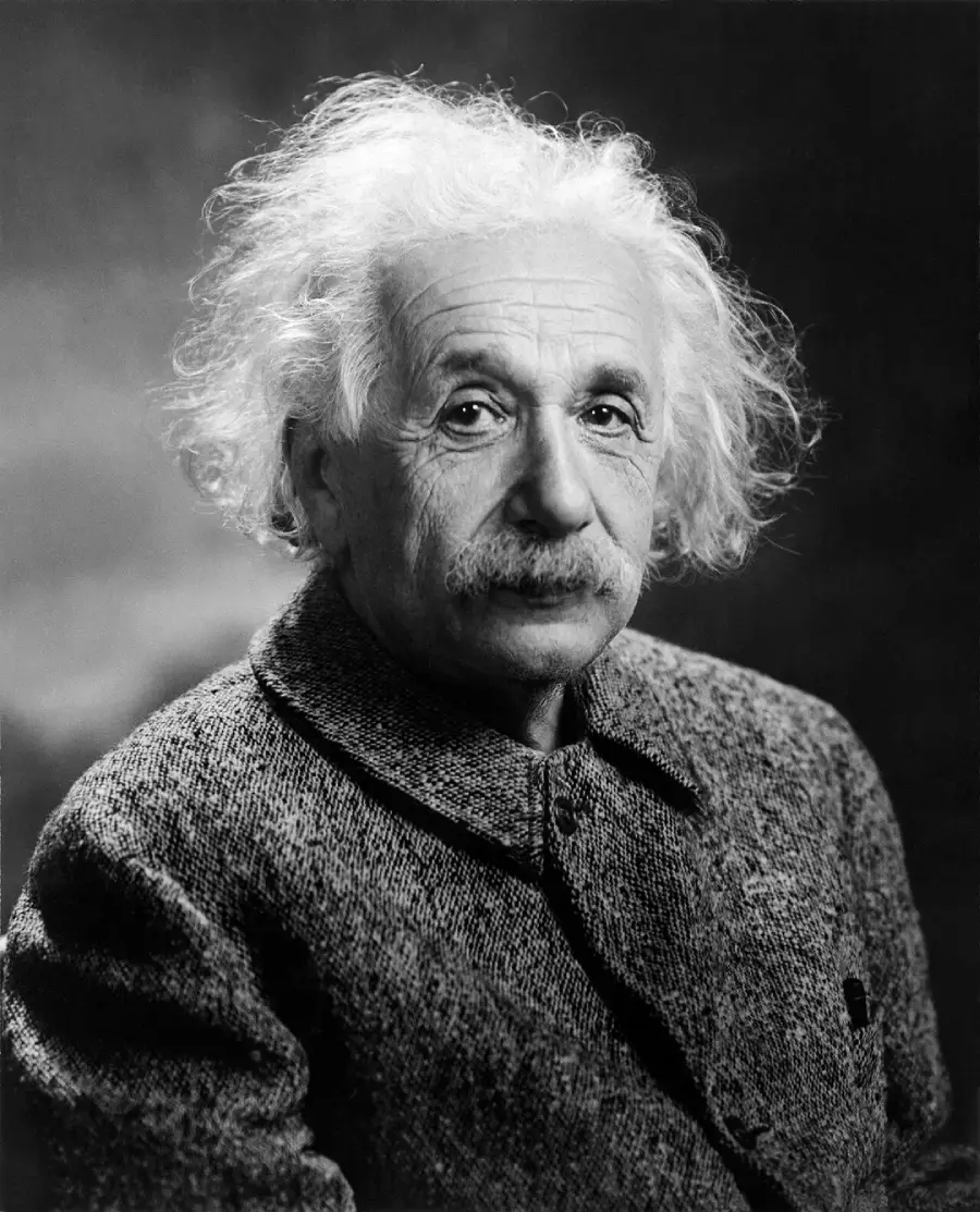 A black-and-white portrait of an older man with wild, white hair and a mustache, wearing a buttoned-up tweed coat, looking calmly at the camera against a plain background.