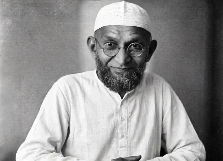 An elderly man with a gray beard, wearing round glasses, a white cap, and a white traditional shirt, poses in front of a plain background.