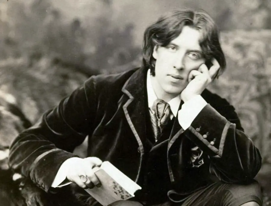 A person with medium-length hair in vintage clothing sits, resting their face on one hand and holding a book in the other, gazing thoughtfully at the camera.