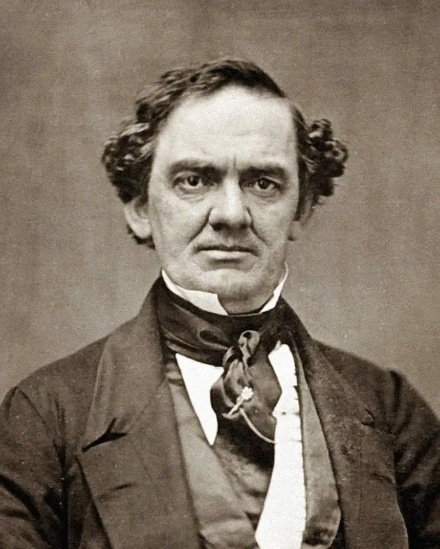 Black and white portrait of a middle-aged man with wavy hair, wearing a dark suit, white shirt, vest, and a cravat tied at the neck, looking directly at the camera with a serious expression.