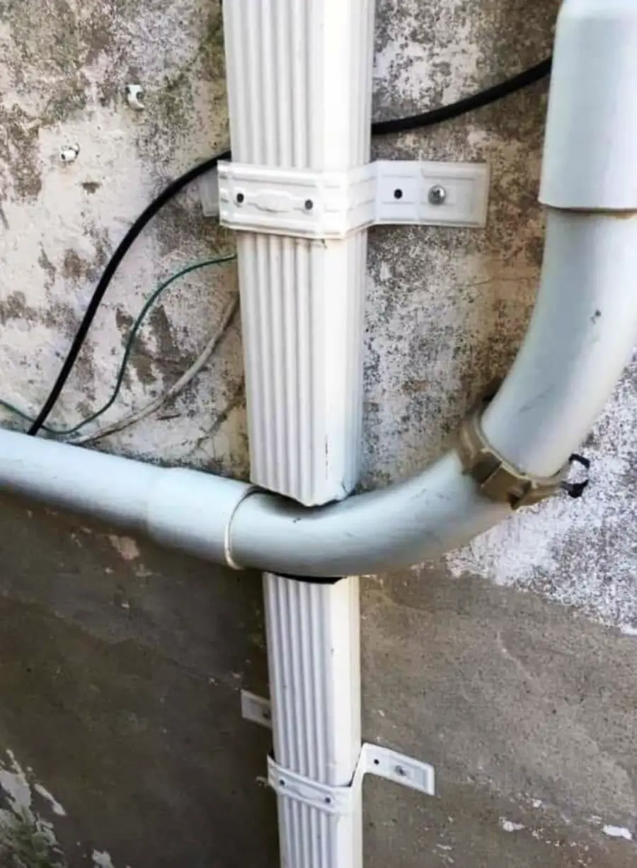 White gutter pipes are awkwardly installed on a weathered wall, with a vertical segment interrupted by a horizontal pipe passing directly through it.