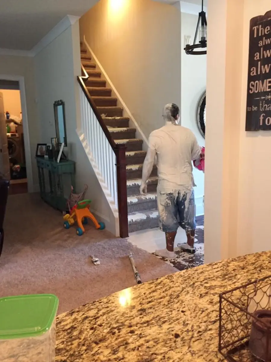 A man covered in white paint stands near stairs with spilled paint on them and the carpet. A fallen paint can and roller lie nearby. The scene looks messy and chaotic.