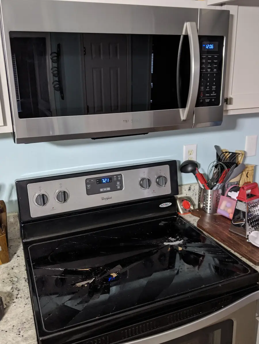 A kitchen stove with a shattered glass cooktop sits beneath a microwave. Utensils and kitchen items are on the counter to the right. The microwave display shows 2:06, and the oven display shows 12:11.