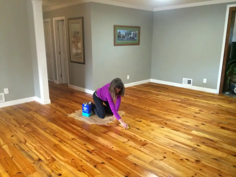 A woman in a purple shirt kneels on a wooden floor, applying a cleaning or polishing product with a sponge in a mostly empty, gray-walled room.