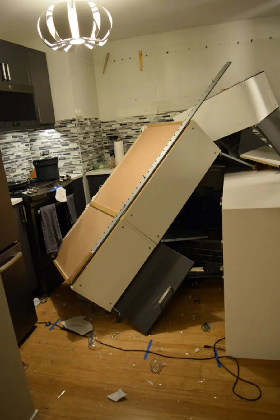 Kitchen scene with a large cabinet that has fallen off the wall, landing on the counter and floor. Broken glass and debris are scattered around, and appliances are partially visible amid the mess.
