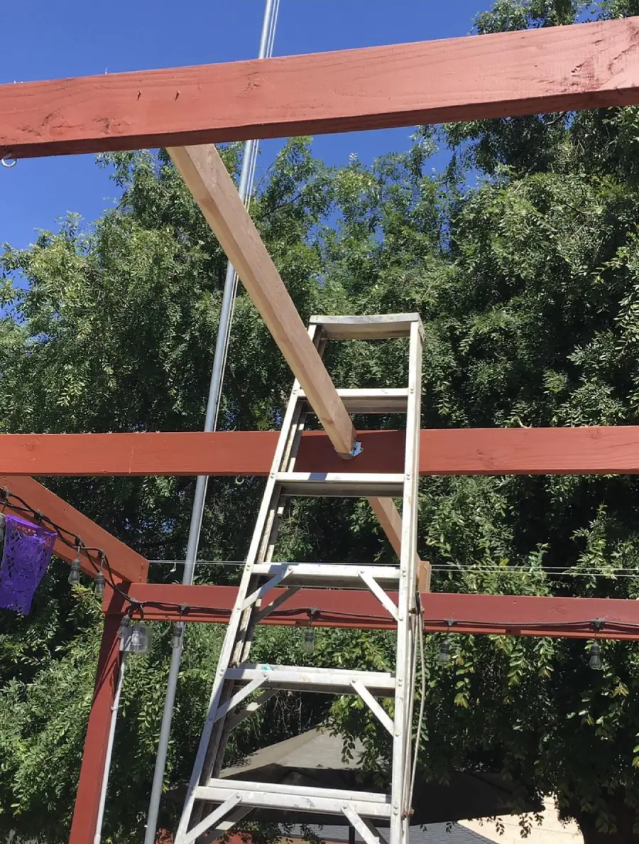 A ladder is precariously balanced on a wooden beam, wedged between two red beams outdoors with trees and a blue sky in the background. The setup appears unsafe.