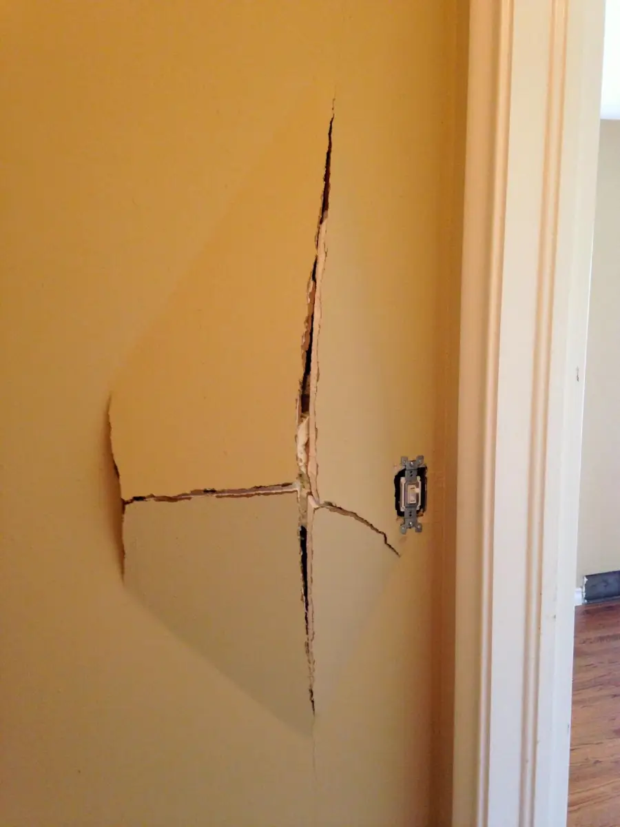 A beige wall with a large, jagged crack in the shape of an uneven cross near a light switch, exposing white material underneath.