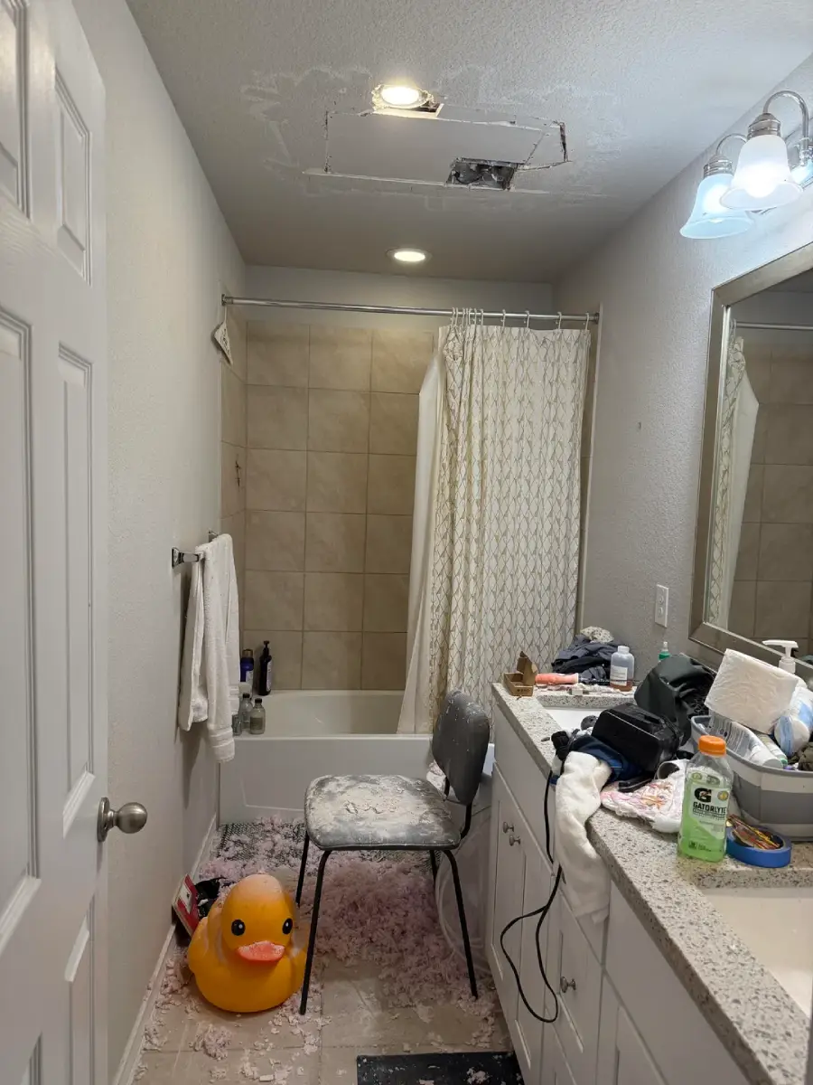 A bathroom with a messy counter, a shower, a towel on the rack, a chair covered in dust, a rubber duck in the tub, pink debris on the floor, and a hole in the ceiling.