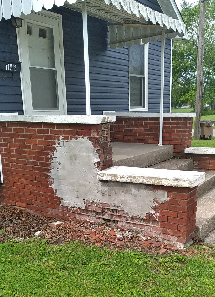 A brick porch has a large section covered in gray cement patchwork; loose bricks and debris are scattered on the grass below.