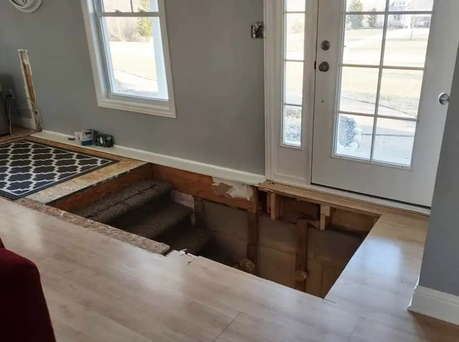 A room with light wood flooring has a large hole exposing stairs and wood framing near double doors, making the area unsafe.
