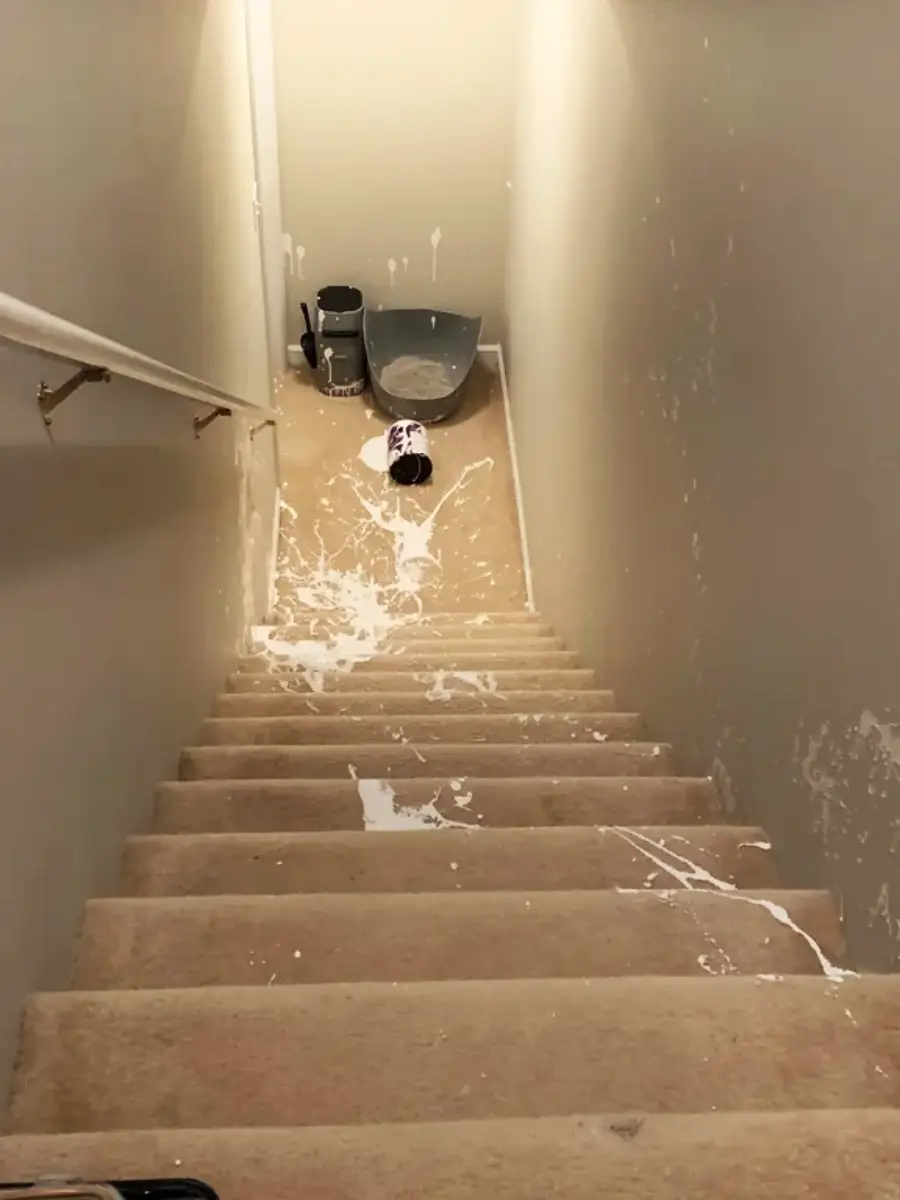 A staircase with beige carpet covered in spilled white paint, leading to a knocked-over paint can, roller tray, and painting supplies at the bottom of the stairs.