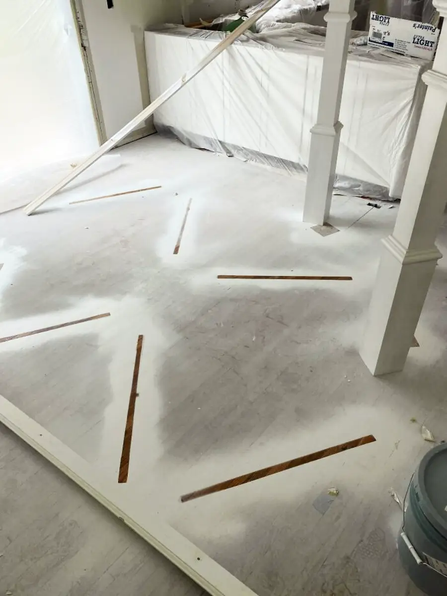 A room under renovation with white dust covering the floor, wooden slats partially visible, and furniture covered by plastic sheets in the background. Painting supplies and construction materials are scattered around.