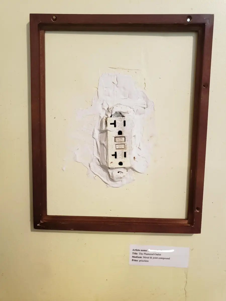 A framed power outlet with messy plaster around it hangs on a wall like artwork. A small label below mimics a museum art description.