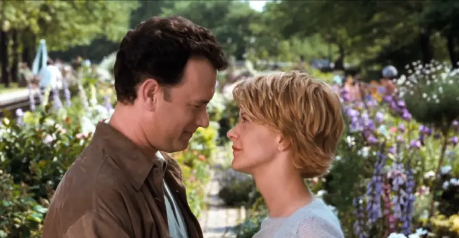 A man and woman stand face to face in a garden filled with colorful flowers, gazing into each other's eyes with gentle smiles. Trees and blurred figures are visible in the background.
