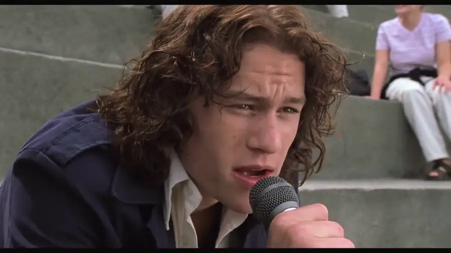 A young man with wavy brown hair sings into a microphone while sitting on outdoor concrete steps, with a blurred person in the background.