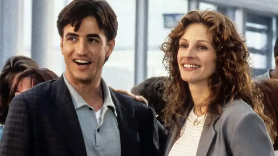 A man and a woman stand close together, smiling, in a crowded indoor setting. The woman has wavy brown hair and the man has short dark hair; both are dressed in casual business attire.