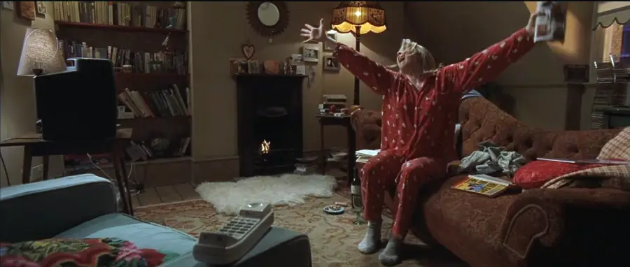 A woman in red pajamas sits on a brown sofa with her arms raised, celebrating while watching TV in a cozy, cluttered living room with warm lighting and bookshelves. A phone rests on the arm of a nearby chair.