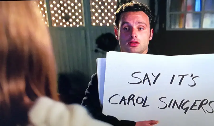 A man stands outside at night holding a sign that reads "SAY IT'S CAROL SINGERS" to a woman whose back is to the camera. String lights are visible in the background.