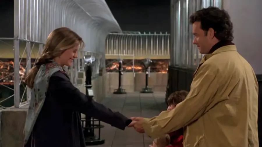 A man and woman smile at each other and hold hands on an observation deck at night; a child is partially visible between them. The setting appears calm and sentimental.