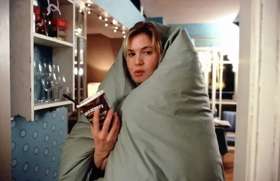 A woman wrapped in a large blanket holds a tub of ice cream and a spoon, standing in a cozy, softly lit room. She looks thoughtful and slightly disheveled.